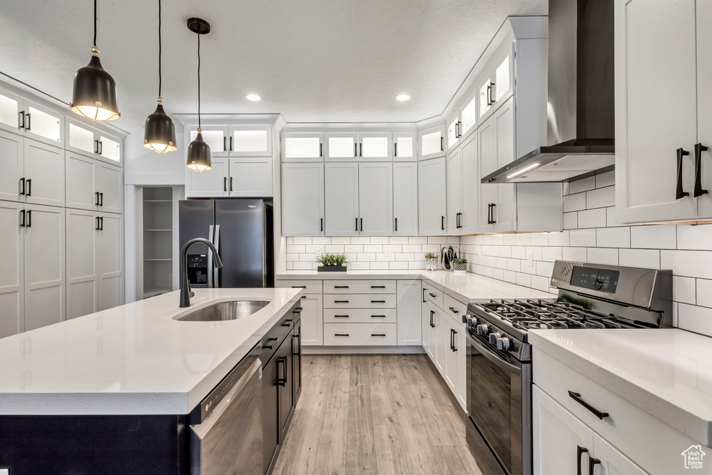 Kitchen featuring wall chimney range hood, light wood-type flooring, white cabinetry, stainless steel appliances, and pendant lighting