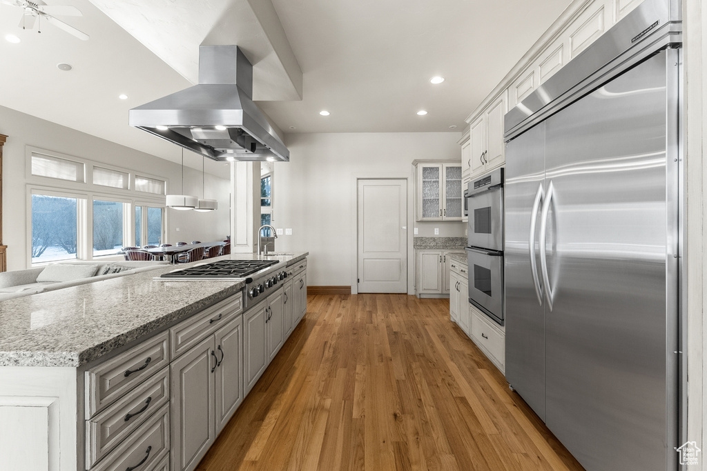 Kitchen with light stone counters, appliances with stainless steel finishes, white cabinetry, island exhaust hood, and light hardwood / wood-style flooring
