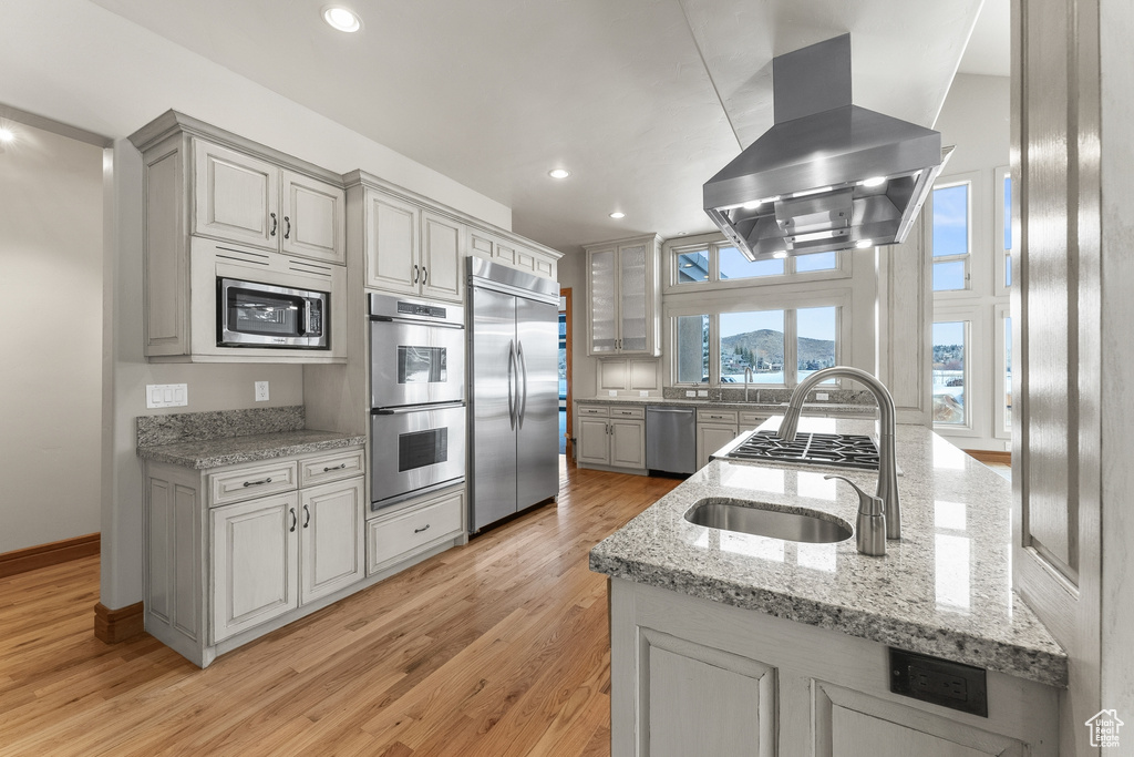 Kitchen with built in appliances, light stone countertops, island range hood, sink, and light hardwood / wood-style floors