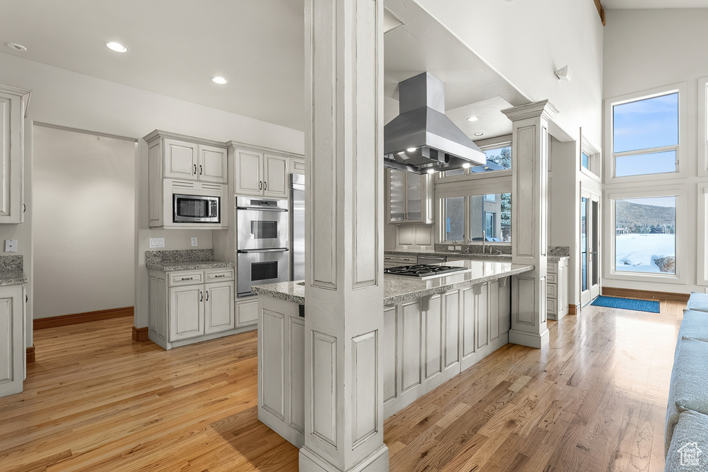 Kitchen with high vaulted ceiling, light hardwood / wood-style flooring, island range hood, stainless steel appliances, and light stone counters