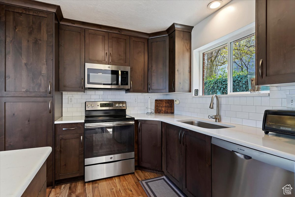 Kitchen with sink, backsplash, stainless steel appliances, dark brown cabinetry, and light wood-type flooring