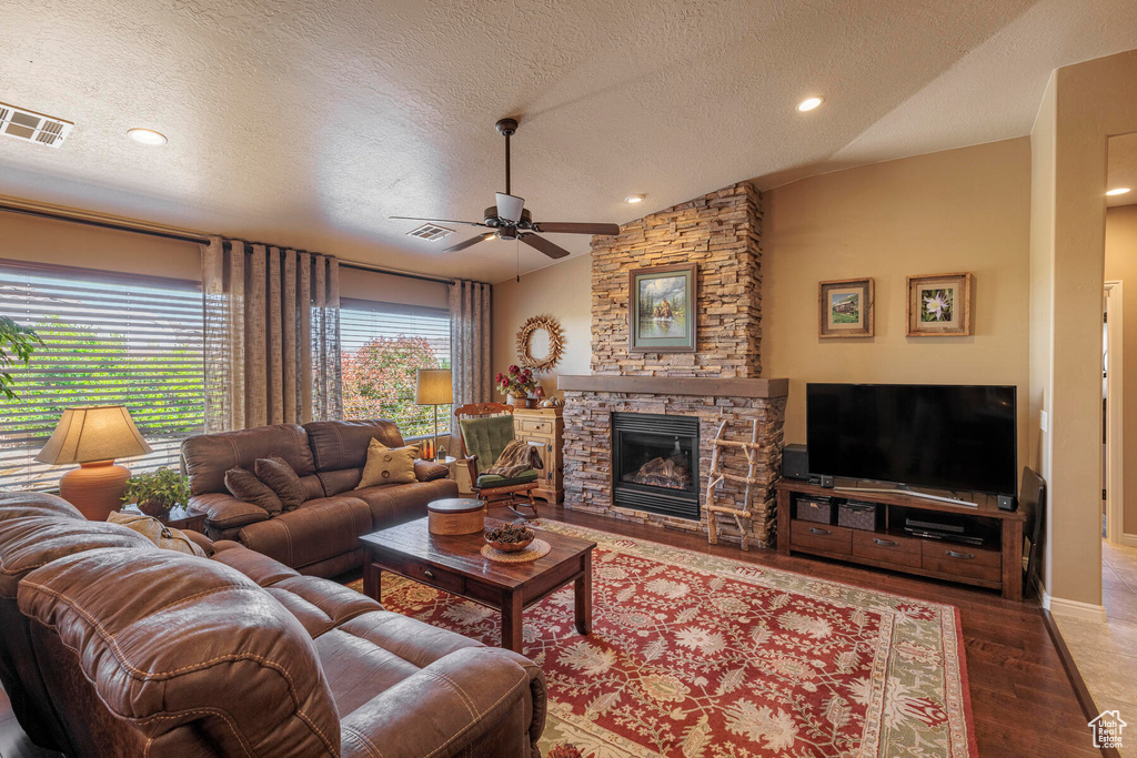 Living room featuring vaulted ceiling, hardwood / wood-style flooring, a stone fireplace, ceiling fan, and a textured ceiling