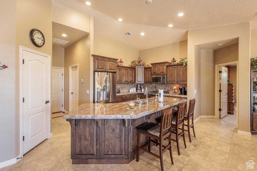 Kitchen with backsplash, stainless steel appliances, an island with sink, a kitchen breakfast bar, and light stone countertops