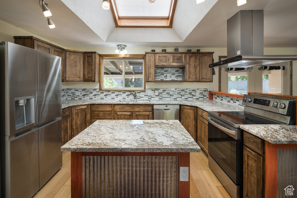Kitchen with a skylight, light stone counters, backsplash, stainless steel appliances, and island exhaust hood