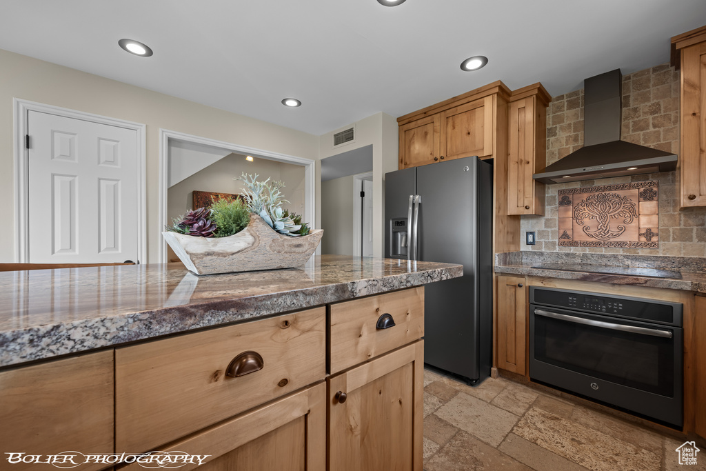 Kitchen featuring appliances with stainless steel finishes, wall chimney exhaust hood, light tile floors, tasteful backsplash, and dark stone counters
