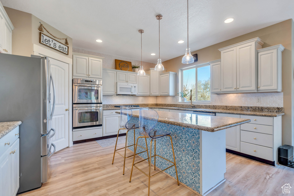 Kitchen featuring appliances with stainless steel finishes, a kitchen island, white cabinetry, decorative light fixtures, and light hardwood / wood-style flooring