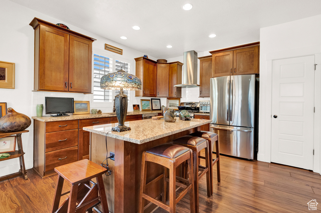 Kitchen featuring dark hardwood / wood-style flooring, wall chimney range hood, light stone counters, a kitchen island, and stainless steel fridge