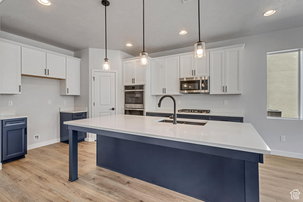 Kitchen featuring white cabinets, decorative light fixtures, stainless steel appliances, light hardwood / wood-style flooring, and a kitchen island with sink