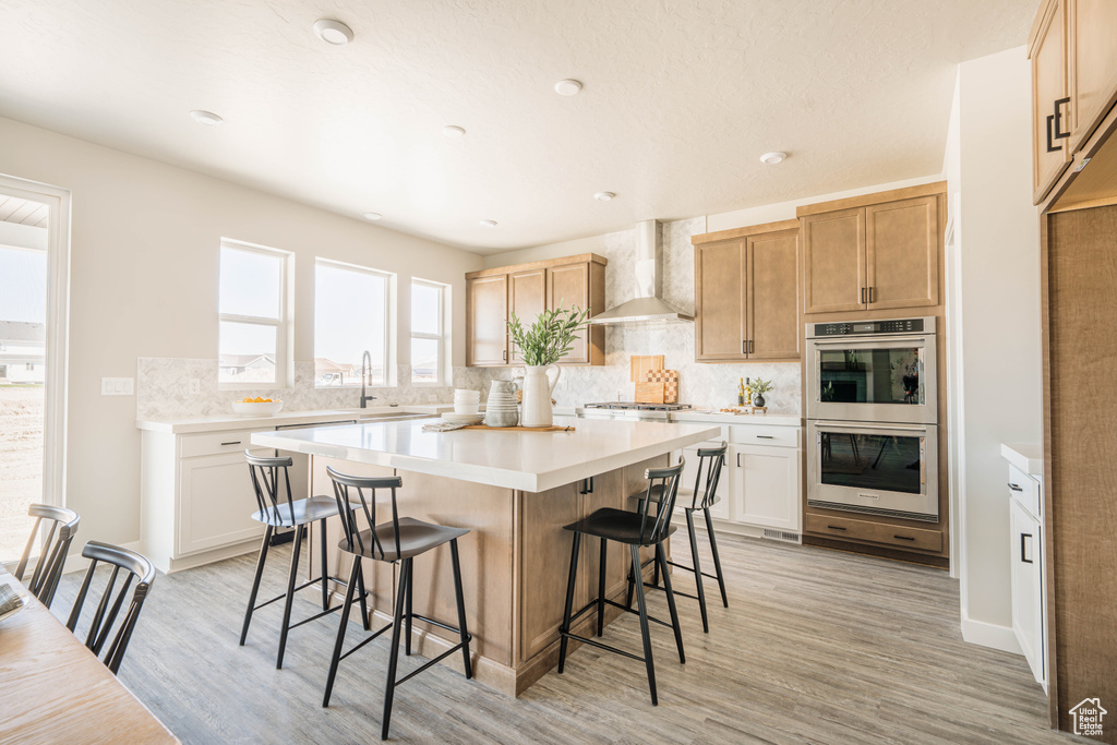Kitchen with appliances with stainless steel finishes, a kitchen island, light hardwood / wood-style floors, wall chimney range hood, and backsplash