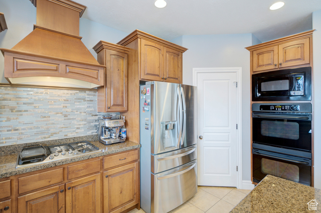 Kitchen with black appliances, backsplash, custom range hood, light tile floors, and light stone counters