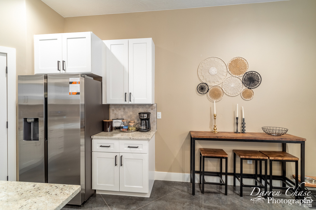 Kitchen with tasteful backsplash, white cabinets, light stone countertops, stainless steel fridge with ice dispenser, and dark tile flooring