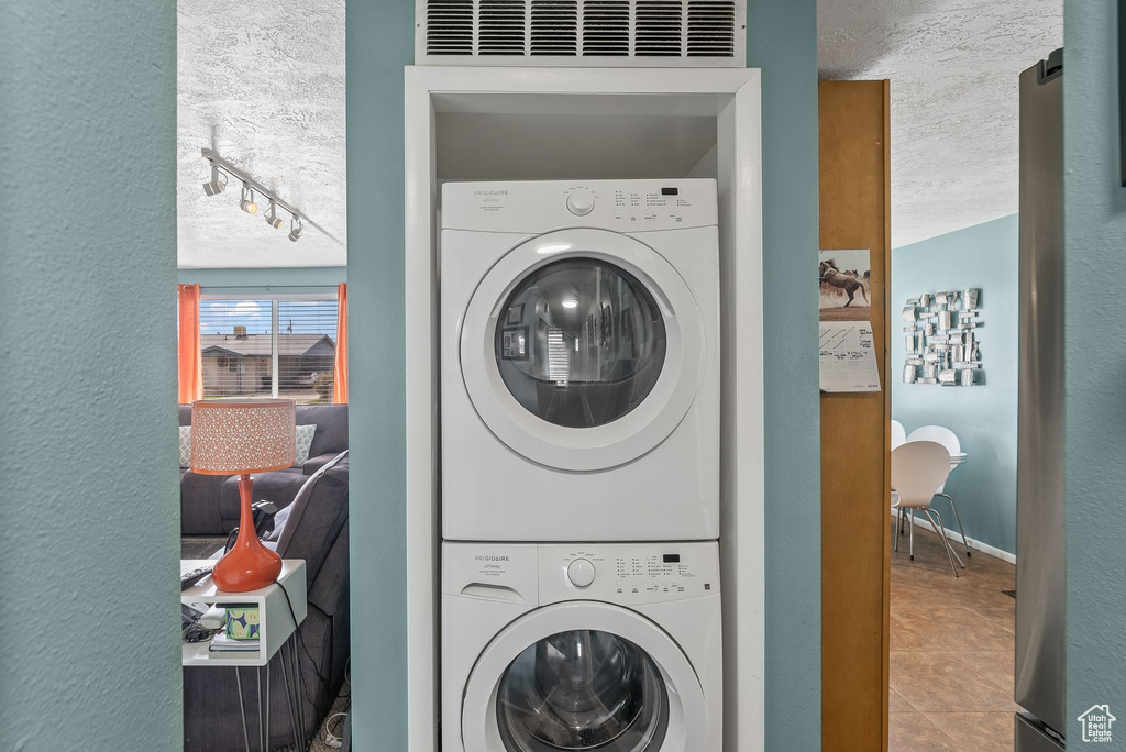 Laundry room featuring track lighting, a textured ceiling, stacked washing maching and dryer, and light tile floors