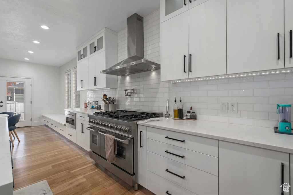 Kitchen featuring backsplash, light hardwood / wood-style flooring, stainless steel appliances, wall chimney range hood, and white cabinets