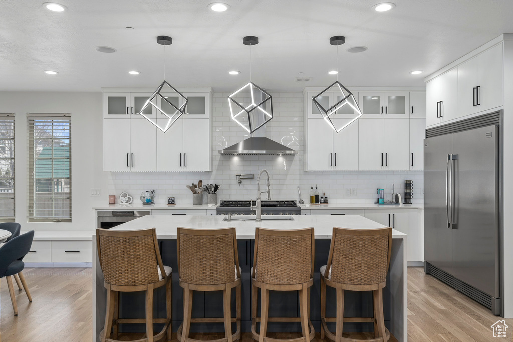 Kitchen featuring wall chimney range hood, stainless steel built in fridge, white cabinetry, and light hardwood / wood-style floors