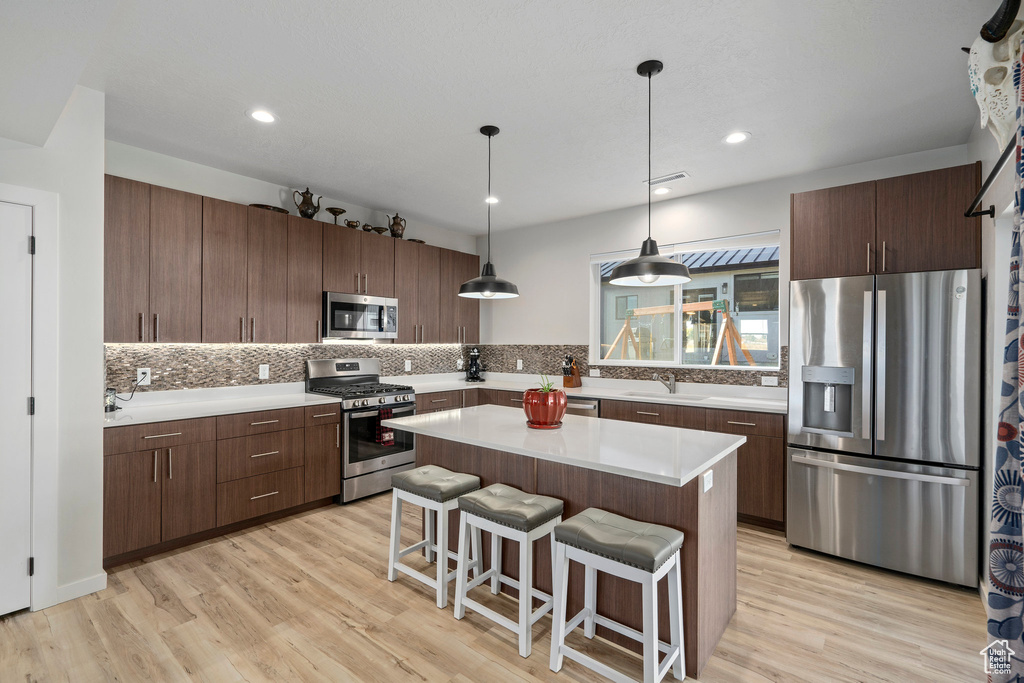 Kitchen featuring appliances with stainless steel finishes, a kitchen island, tasteful backsplash, hanging light fixtures, and light hardwood / wood-style flooring