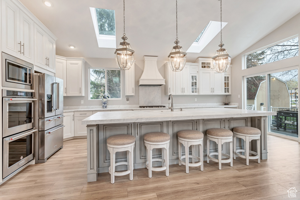 Kitchen featuring vaulted ceiling with skylight, stainless steel appliances, a center island with sink, premium range hood, and pendant lighting