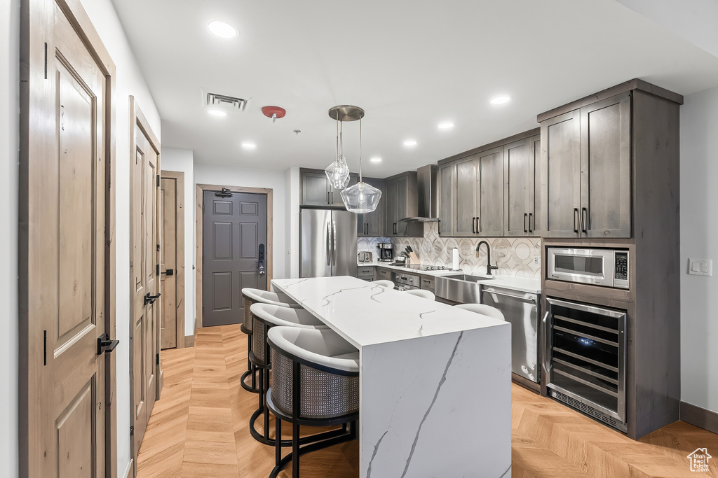 Kitchen with light parquet floors, stainless steel appliances, wine cooler, wall chimney range hood, and pendant lighting