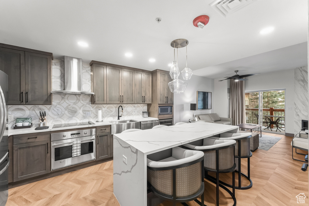 Kitchen featuring appliances with stainless steel finishes, ceiling fan, wall chimney exhaust hood, light parquet flooring, and hanging light fixtures