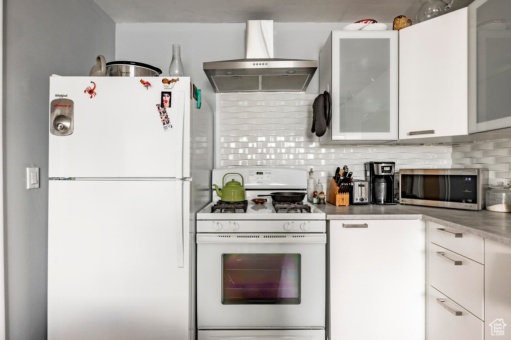 Kitchen with white appliances, wall chimney range hood, tasteful backsplash, and white cabinetry
