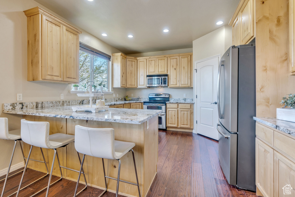 Kitchen featuring light brown cabinets, light stone countertops, appliances with stainless steel finishes, a breakfast bar area, and dark hardwood / wood-style floors