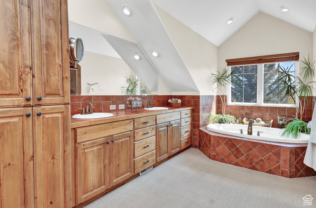 Bathroom with tile walls, vaulted ceiling, dual bowl vanity, and tiled bath