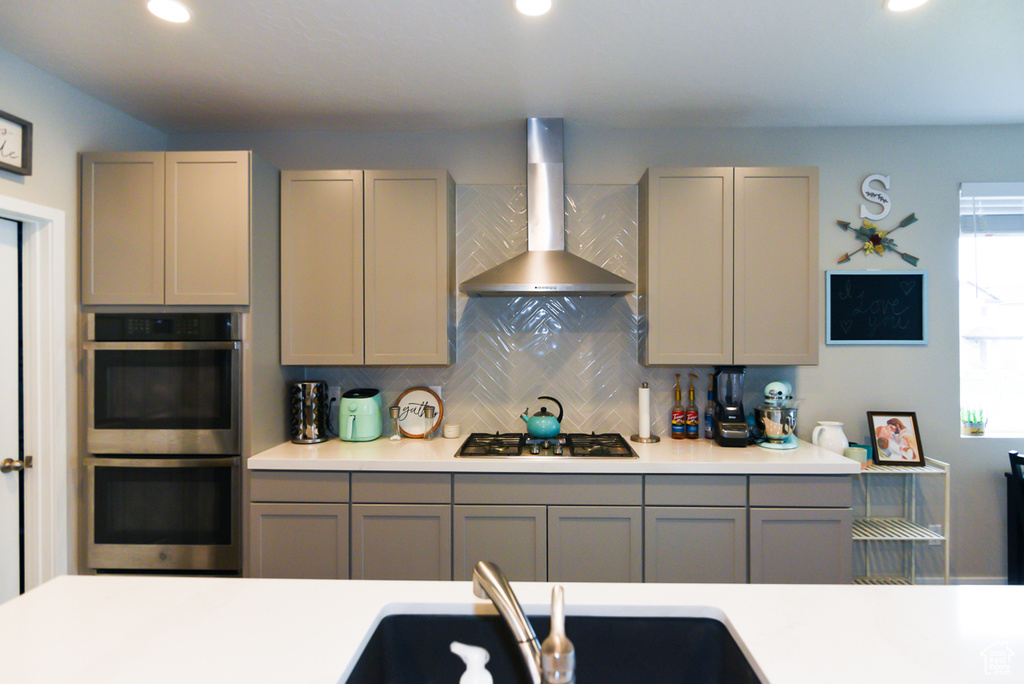 Kitchen featuring backsplash, gray cabinets, double oven, gas stovetop, and wall chimney range hood