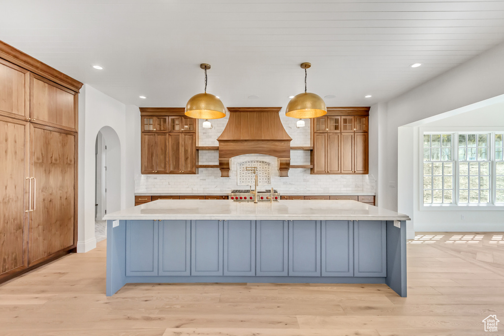 Kitchen with backsplash, premium range hood, light hardwood / wood-style flooring, and a kitchen island with sink
