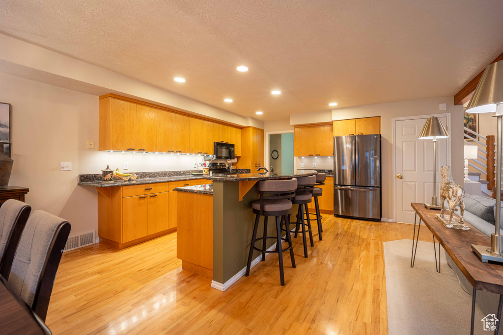 Kitchen with a kitchen island, a breakfast bar, stainless steel refrigerator, dark stone counters, and light hardwood / wood-style flooring
