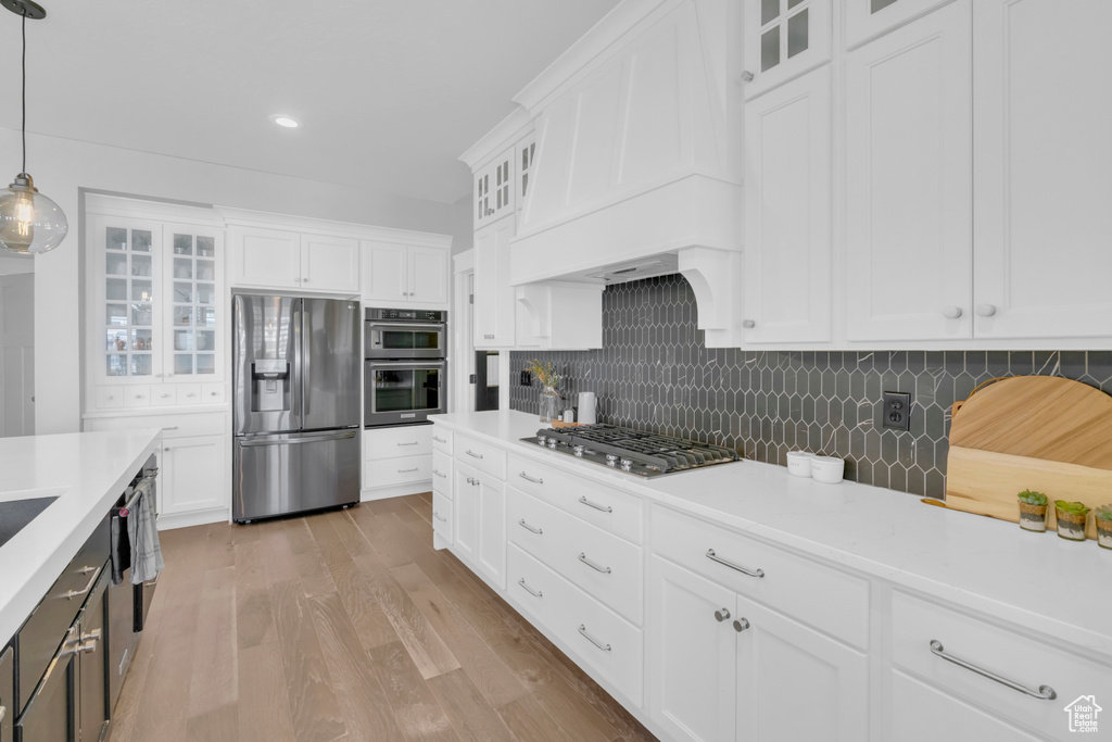 Kitchen with hanging light fixtures, white cabinetry, appliances with stainless steel finishes, custom range hood, and light hardwood / wood-style floors