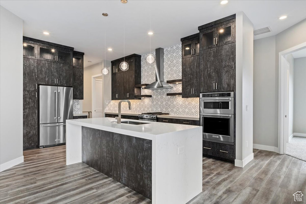 Kitchen with decorative light fixtures, appliances with stainless steel finishes, light hardwood / wood-style flooring, wall chimney exhaust hood, and a kitchen island with sink