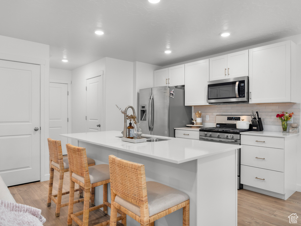 Kitchen featuring appliances with stainless steel finishes, light hardwood / wood-style flooring, a kitchen island with sink, white cabinetry, and a kitchen breakfast bar