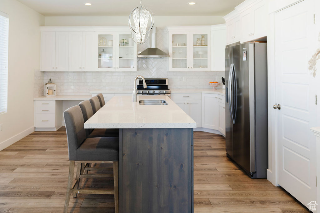 Kitchen featuring light wood-type flooring, wall chimney range hood, stainless steel appliances, a center island with sink, and tasteful backsplash