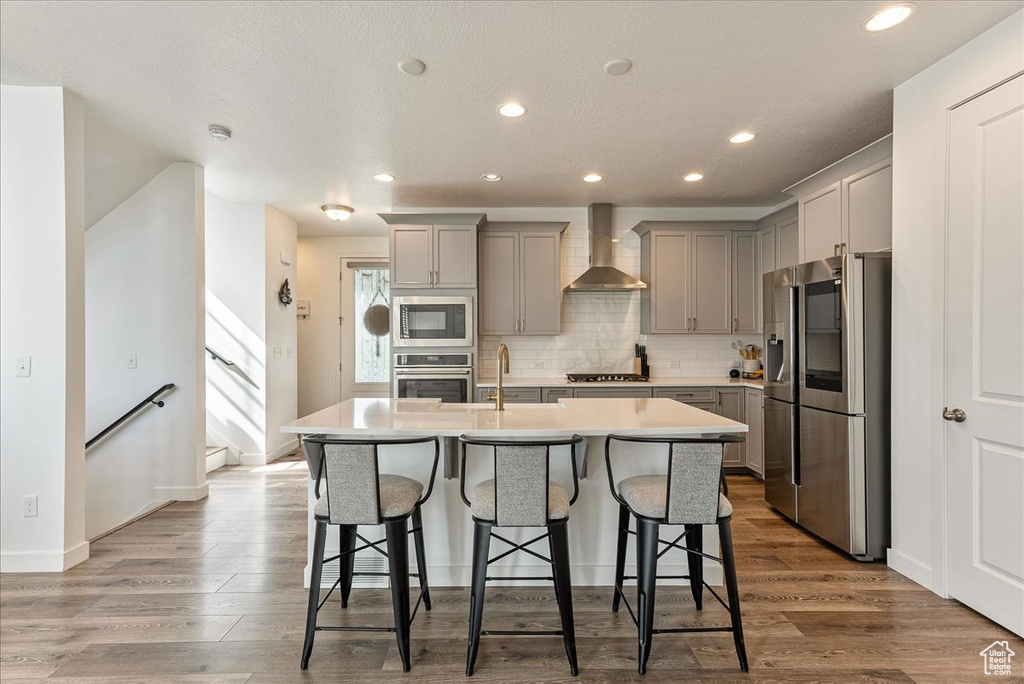 Kitchen featuring appliances with stainless steel finishes, wall chimney range hood, a kitchen island with sink, and light hardwood / wood-style floors