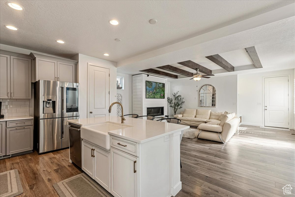 Kitchen featuring beamed ceiling, hardwood / wood-style floors, appliances with stainless steel finishes, a center island with sink, and ceiling fan