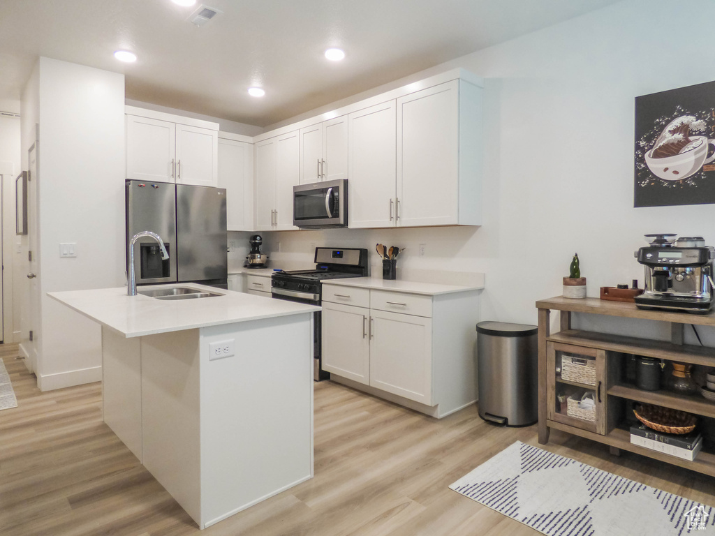 Kitchen with appliances with stainless steel finishes, light hardwood / wood-style floors, white cabinets, and an island with sink