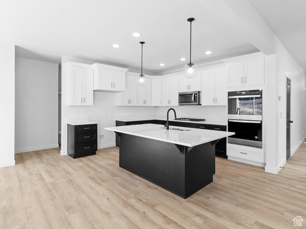 Kitchen featuring white cabinets, light hardwood / wood-style floors, decorative light fixtures, stainless steel appliances, and a kitchen island with sink