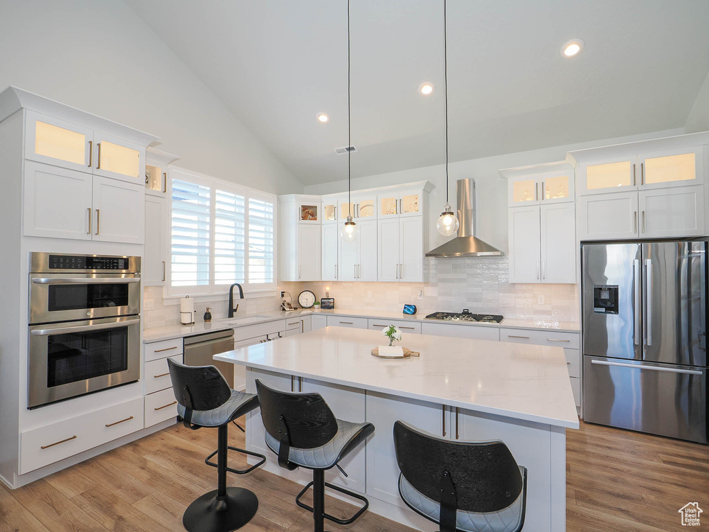 Kitchen featuring wall chimney exhaust hood, hanging light fixtures, appliances with stainless steel finishes, a kitchen island, and tasteful backsplash