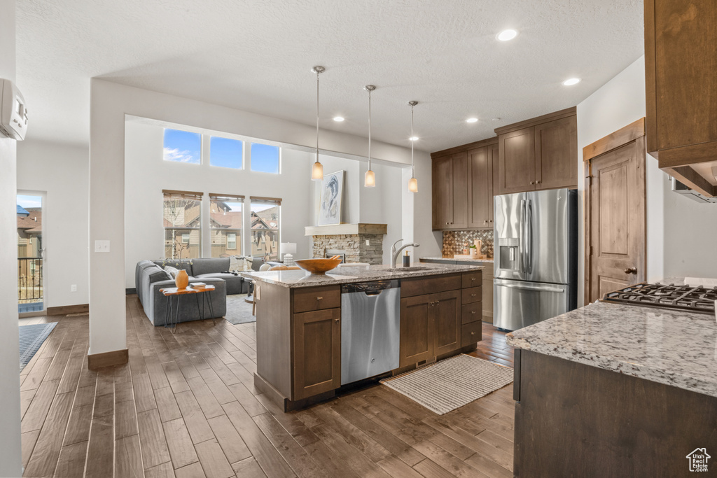 Kitchen featuring light stone counters, tasteful backsplash, decorative light fixtures, stainless steel appliances, and a kitchen island with sink