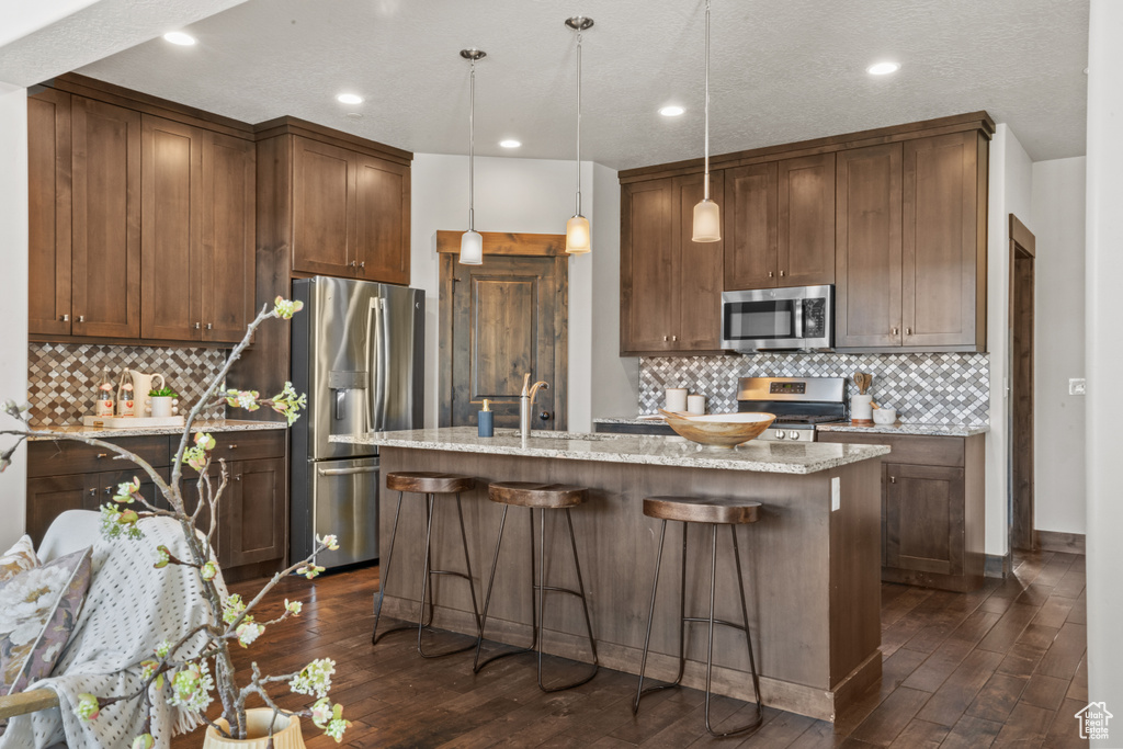 Kitchen featuring a kitchen island with sink, hanging light fixtures, appliances with stainless steel finishes, light stone counters, and tasteful backsplash