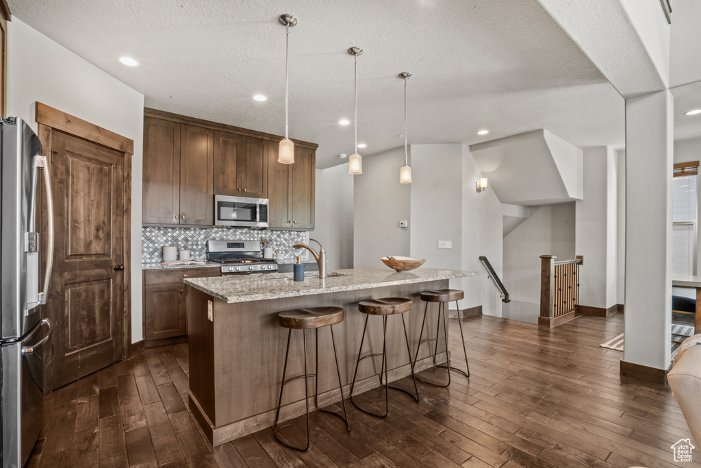 Kitchen with appliances with stainless steel finishes, dark hardwood / wood-style floors, decorative light fixtures, and a kitchen island with sink