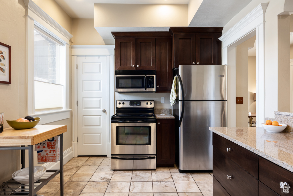 Kitchen with dark brown cabinetry, stainless steel appliances, light stone countertops, and light tile flooring