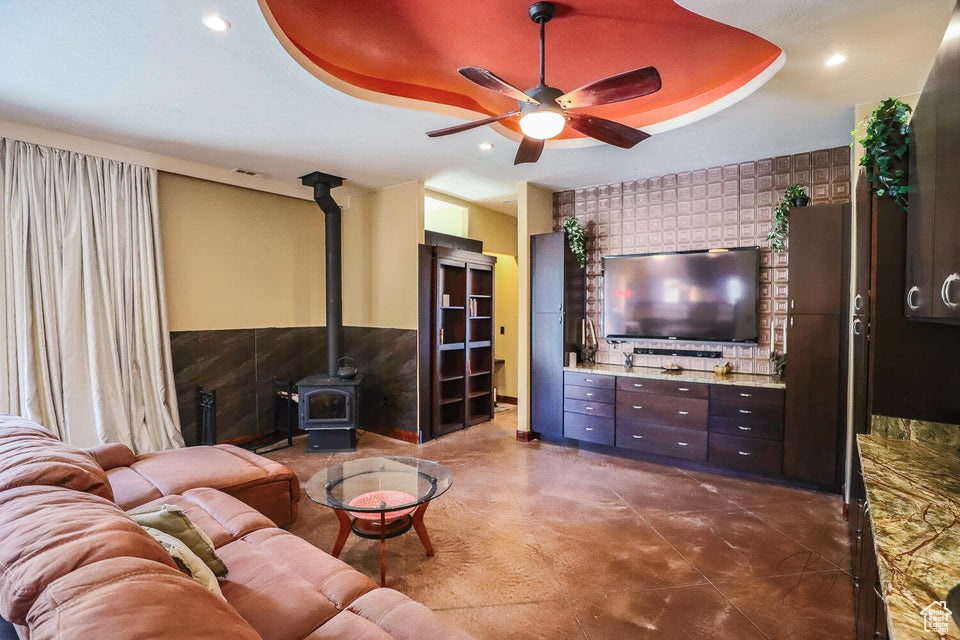 Living room featuring tile walls, a wood stove, ceiling fan, and a tray ceiling