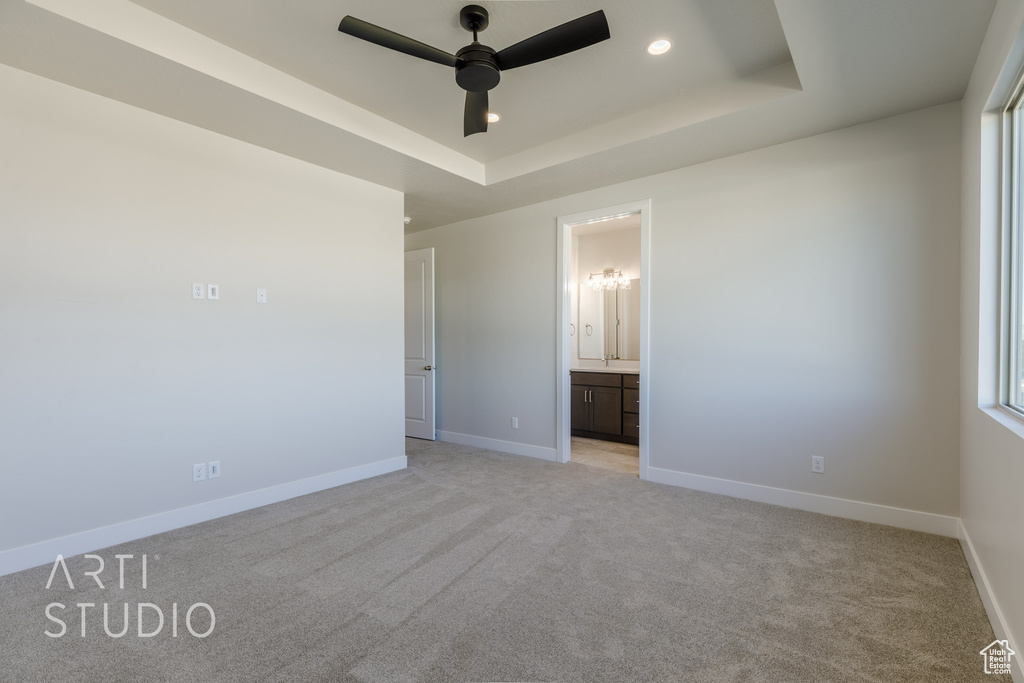 Unfurnished bedroom featuring ensuite bath, a tray ceiling, light colored carpet, and ceiling fan