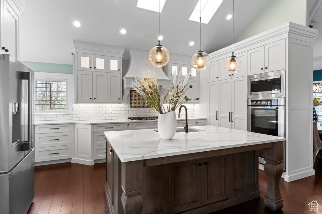 Kitchen featuring appliances with stainless steel finishes, hanging light fixtures, backsplash, dark wood-type flooring, and a kitchen island with sink