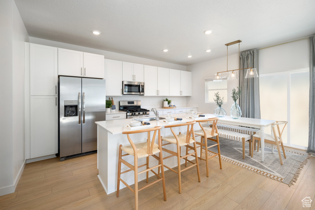 Kitchen featuring appliances with stainless steel finishes, light wood-type flooring, white cabinetry, an island with sink, and pendant lighting
