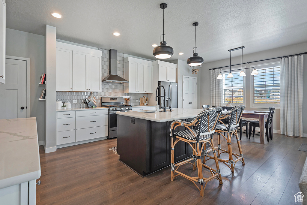 Kitchen with hanging light fixtures, dark hardwood / wood-style flooring, appliances with stainless steel finishes, wall chimney range hood, and a center island with sink
