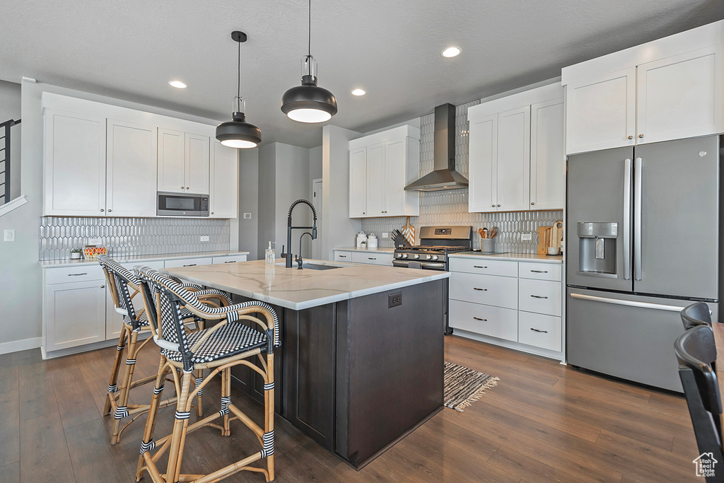 Kitchen with wall chimney exhaust hood, a kitchen island with sink, dark wood-type flooring, backsplash, and appliances with stainless steel finishes