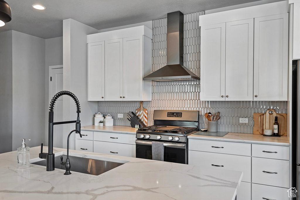 Kitchen featuring gas stove, wall chimney exhaust hood, white cabinetry, and light stone counters