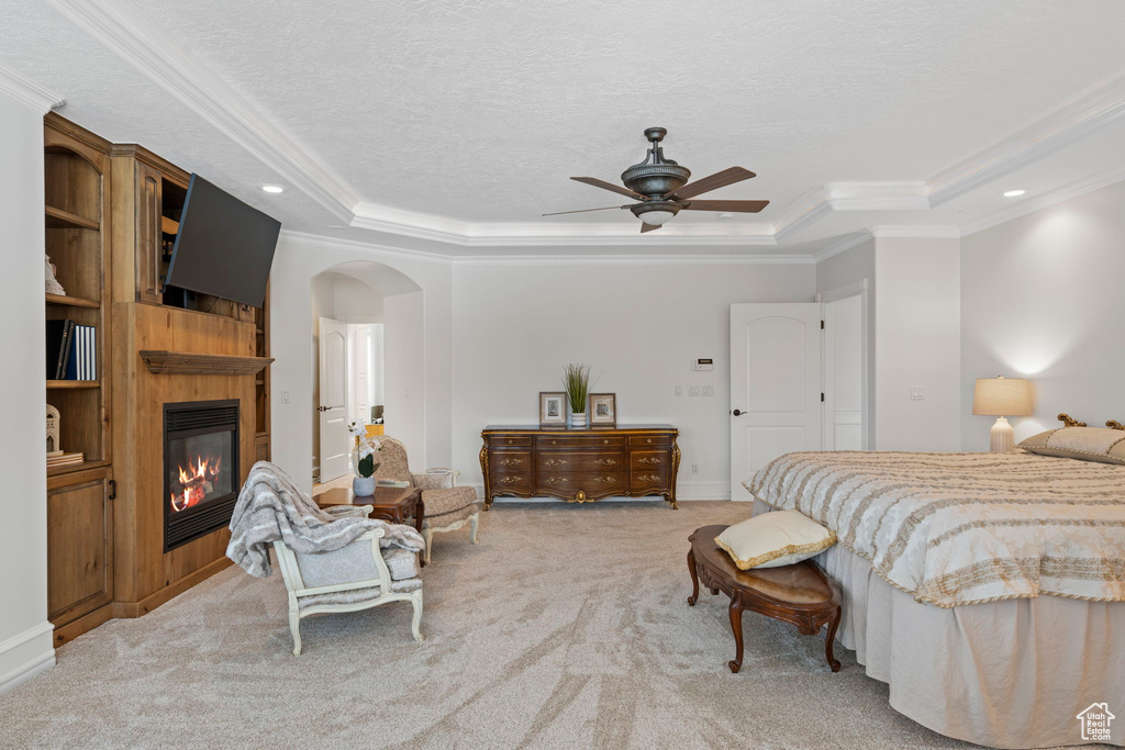 Bedroom with ceiling fan, a fireplace, a raised ceiling, light colored carpet, and crown molding