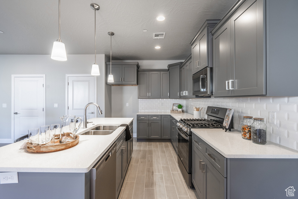 Kitchen featuring sink, appliances with stainless steel finishes, hanging light fixtures, a center island with sink, and gray cabinetry